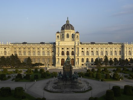 Das Bild zeigt die prächtige Fassade des Kunsthistorischen Museums in Wien, umgeben von gepflegten Gärten und Bäumen. Im Vordergrund steht ein Denkmal, das die Aufmerksamkeit auf die imposante Architektur und den geschmackvollen Landschaftsgestaltung lenkt.