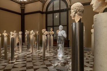 Eine Galerie mit mehreren Skulpturen und Büsten, die auf Säulen stehen. In der Mitte befindet sich eine stilisierte, transparente Figur. Der Fußboden ist schachbrettartig weiß und dunkel, während hohe Fenster das Licht hereinlassen.