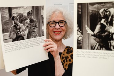 Eine lächelnde Frau mit Brille hält zwei große Bilder mit historischen Fotografien in der Hand. Im Hintergrund sind unscharfe Details einer Ausstellung zu erkennen. Die Frau trägt ein Oberteil mit einem Leopardenmuster.