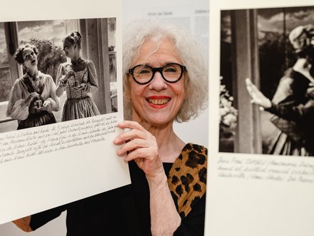 Eine lächelnde Frau mit Brille hält zwei große Bilder mit historischen Fotografien in der Hand. Im Hintergrund sind unscharfe Details einer Ausstellung zu erkennen. Die Frau trägt ein Oberteil mit einem Leopardenmuster.