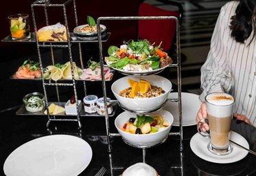 Ein stilvolles Buffet mit mehreren Ebenen: Oben sind kleine Desserts und Kekse, in der Mitte eine große Schüssel mit Salat, darunter eine Schüssel mit Müsli und frischen Früchten. Auf der Seite steht ein Glas Kaffee. Der Tisch ist elegant gedeckt.