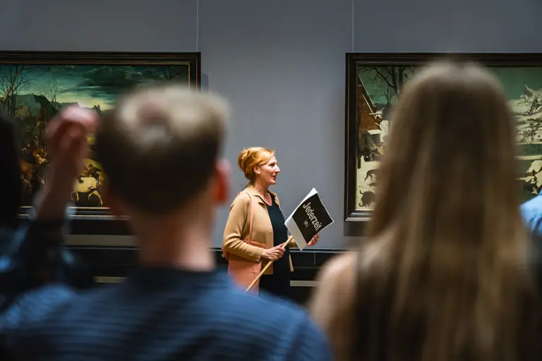 Eine Museumsguides steht inmitten eines Raumes mit zwei Gemälden im Hintergrund. Sie hält ein Schild mit dem Wort 