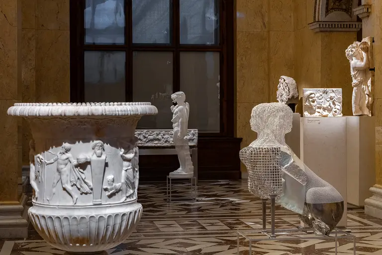 Eine Kunstinstallation im Museum zeigt eine weiße Marmorschale mit Reliefs von Figuren, eine skulpturierte menschliche Figur und eine durchsichtige, abstrahierte Figur. Die Werke sind auf Sockeln platziert und vor einem fensterähnlichen Hintergrund aus Holz.