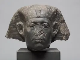Eine antike ägyptische Statue eines Pharaos, hauptsächlich der Kopf in dunklem Stein. Der Kopf hat charakteristische Merkmale wie eine hohe Krone und feine Details im Gesicht. Einige Bereiche sind beschädigt, aber die ausdrucksvolle Darstellung bleibt erkennbar.