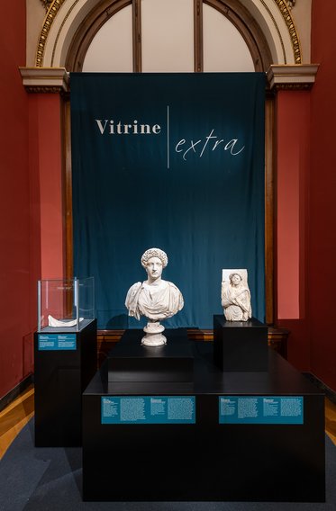 Drei Skulpturen stehen in einer Vitrine in einem Museum. Im Hintergrund hängt ein Banner mit der Aufschrift „Vitrine extra“. Die Skulpturen sind in verschiedenen Größen und Stilen, mit einer Büste in der Mitte und weiteren Stücken auf den Seiten.
