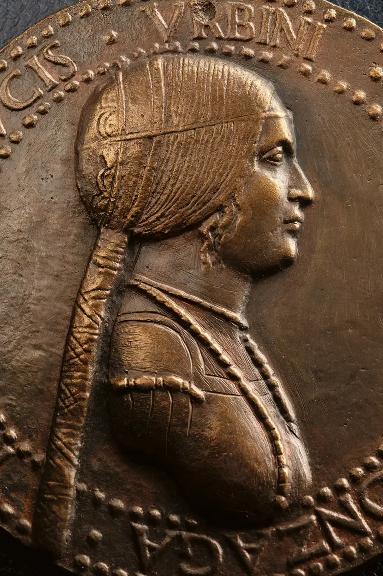 A bronze medal depicts a profile of a woman wearing a detailed headdress and ornate clothing, highlighting her graceful features and jewelry. The inscription around the edge includes a name and possibly a title, suggesting historical significance.
