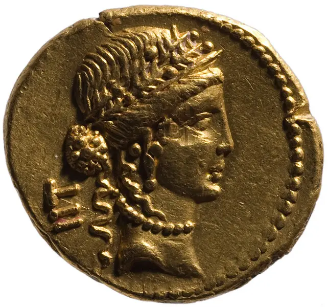 Eine goldene Münze zeigt ein Profilporträt einer Frau mit einer diademähnlichen Krone und lockigem Haar. Die Münze ist detailliert und zeigt feine Linien um das Gesicht und den Hals. Sie stammt aus der Antike und symbolisiert möglicherweise einen Herrscher oder eine Gottheit.