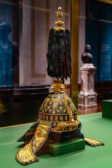 Die Abbildung zeigt eine prunkvolle Kopfbedeckung mit goldenen Verzierungen und einem langen, schwarzen Federbusch. Der untere Teil ist kunstvoll gestaltet, mit Details und Stickereien. Die Hutform ist traditionell und bietet einen Blick auf historisches Handwerk.