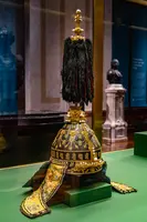 Die Abbildung zeigt eine prunkvolle Kopfbedeckung mit goldenen Verzierungen und einem langen, schwarzen Federbusch. Der untere Teil ist kunstvoll gestaltet, mit Details und Stickereien. Die Hutform ist traditionell und bietet einen Blick auf historisches Handwerk.