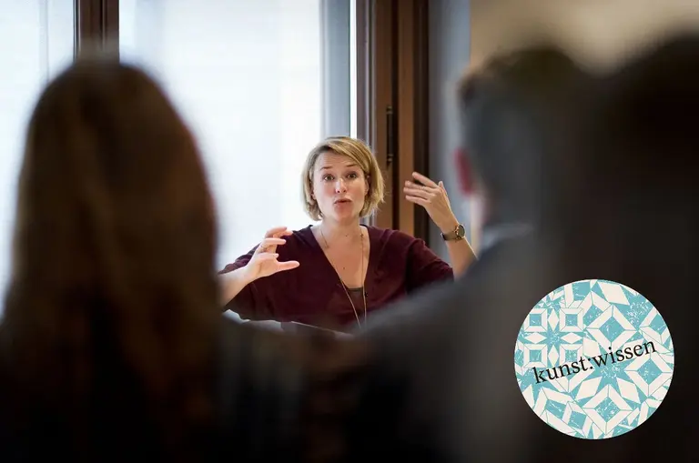 Eine Person spricht lebhaft und gestikuliert in einem Gespräch. Sie sitzt vor einem Fenster und ist von anderen Zuhörern umgeben. Im Vordergrund ist ein Logo mit der Aufschrift „kunst.wissen“ zu sehen.