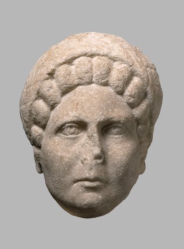 Eine antike Skulptur eines männlichen Kopfes aus Stein, mit detaillierten Zügen, ovale Augen und einer Frisur mit Locken. Der Kopf ist frontal und zeigt eine ernsthafte Miene. Der Hintergrund ist einfarbig und neutral.