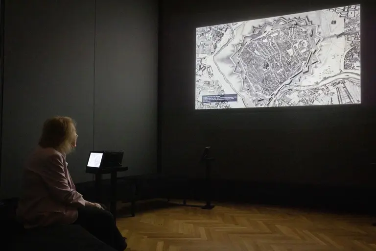 Eine Person sitzt auf einer Bank und betrachtet eine großflächige Projektion einer historischen Stadtkarte an der Wand. Neben der Person steht ein Tisch mit einem Laptop. Die Umgebung ist dunkel und fokussiert auf die Präsentation.