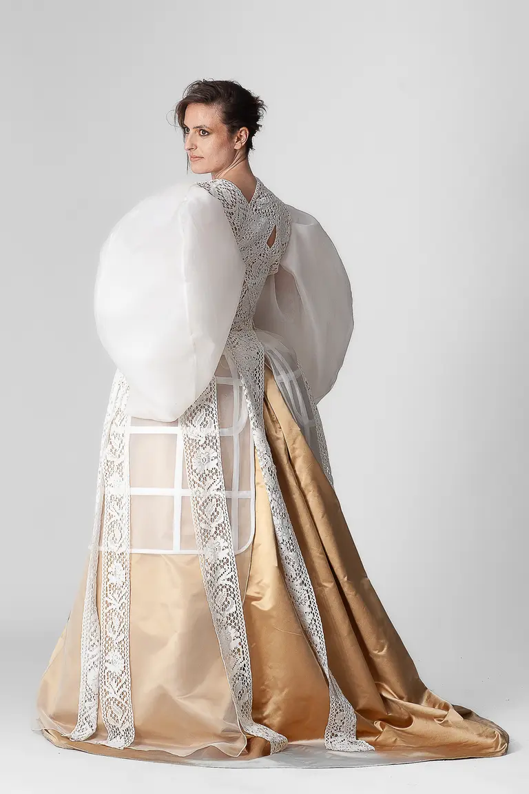 A model poses in a dramatic dress featuring large, billowing white sleeves and a gold satin skirt. The dress has intricate lace detailing and a fitted bodice, showcasing a unique blend of traditional and avant-garde fashion. The background is a simple gray, highlighting the outfit.
