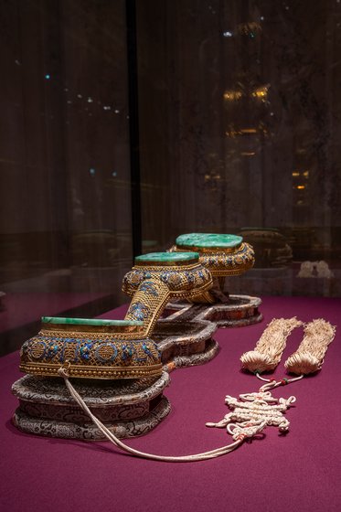 Das Bild zeigt kunstvoll verzierte Schuhmodelle aus der traditionellen Kleidung, die auf einem lila Untergrund liegen. Die Schuhe sind mit bunten Steinen geschmückt und haben eine elegante Form. Daneben stehen weitere dekorative Elemente.