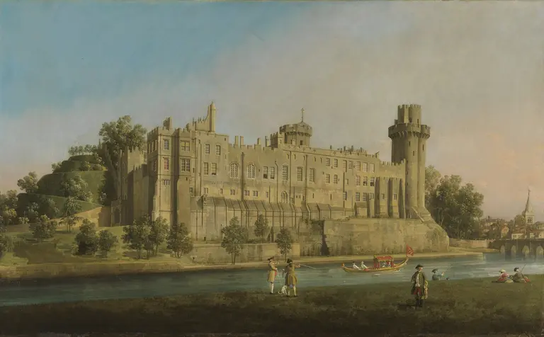 A large, historic castle sits beside a river, surrounded by greenery. In the foreground, people are gathered on the riverbank, some are walking and others are in a boat. The sky is clear, and there are trees and buildings in the background.