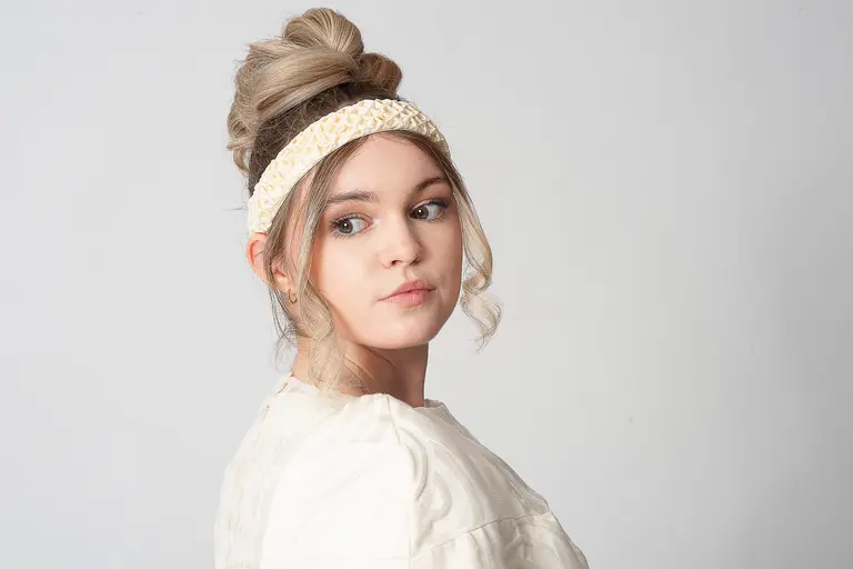 A young woman with a stylish updo and a light-colored headband gazes over her shoulder. She wears a textured cream-colored top and has a thoughtful expression against a neutral background.