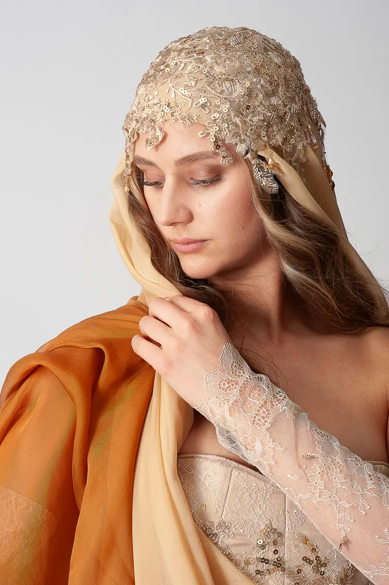 A young woman wears an ornate beaded headpiece and a flowing, sheer shawl. She gazes down thoughtfully, showcasing delicate lace detailing on her arm. The color palette includes warm earth tones, enhancing the elegance of her attire.