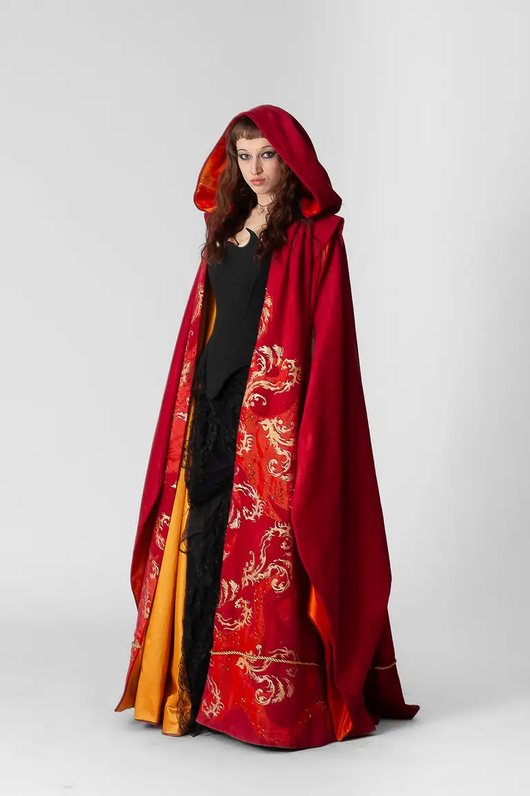 A woman stands in a striking, long red cloak with gold embellishments and a hood. Underneath, she wears a black top and a skirt with intricate details. The background is plain white, allowing the vibrant colors of her outfit to stand out.
