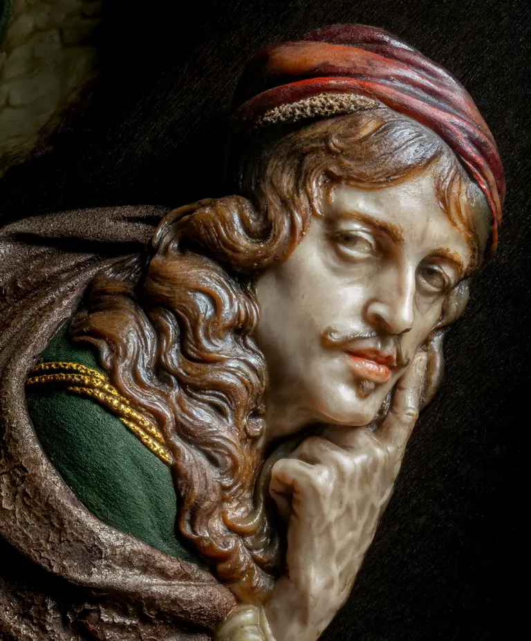 Das Bild zeigt einen nachdenklichen Mann mit langen, lockigen Haaren und einem roten Kopfschmuck. Er legt seinen Zeigefinger an die Lippen, als ob er um Stille bittet. Sein Gesichtsausdruck ist ernst und intensiv, was eine Nachdenklichkeit vermittelt. Er trägt ein grünes Gewand mit goldenen Verzierungen.