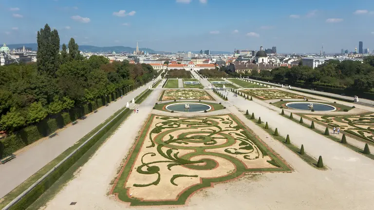 Eine Luftaufnahme eines großen, symmetrischen Gartens mit kunstvollen Beeten, Wasserbecken und geometrischen Mustern. Im Hintergrund sind Stadtgebäude und ein klarer Himmel zu sehen. Der Garten ist von Bäumen und Sträuchern umgeben.