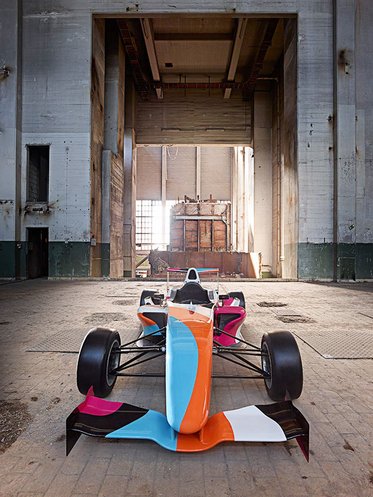 Ein Rennwagen steht in einer weiten, verlassenen Halle. Die Wände sind aus rohem Beton, und es gibt große Fenster, durch die Licht fällt. Der Wagen hat eine auffällige Farbgestaltung in Orange, Blau, Pink und Weiß. Die Szenerie vermittelt einen Eindruck von Verlassenheit und inaktiver Dynamik.