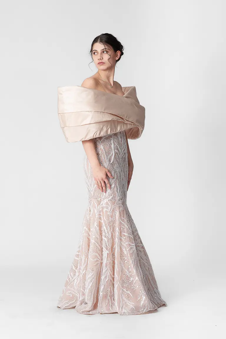 A model poses elegantly in a form-fitting, shimmering gown that features a dramatic, oversized off-shoulder collar. The dress has intricate patterns and a mermaid silhouette, accentuating the model’s figure against a plain white background.