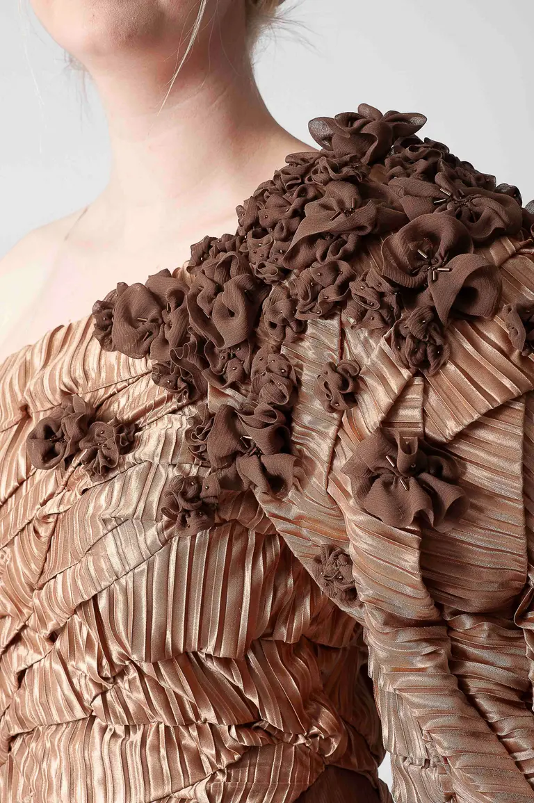A close-up view of a person wearing a elegant, off-shoulder dress featuring a textured, pleated design in a warm, light brown color. The dress is adorned with intricate, fabric floral embellishments on one shoulder, creating a dramatic and artistic effect.