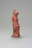 Ein kleines, rotes Figur aus Keramik zeigt eine anthropomorphe Gestalt mit einem Tierkopf. Die Figur steht auf einem transparenten Sockel und hat eine aufrechte Haltung. Sie hält einen Gegenstand in den Händen und hat eine detaillierte Darstellung von Körper und Gesicht.