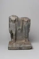 Eine antike Steinskulptur, die zwei menschliche Figuren ohne Köpfe zeigt, die sich an den Händen halten. Die Figur links ist in einem traditionellen Gewand mit kurzer Hose dargestellt, während die rechte Figur in einem langen Gewand zu sehen ist.