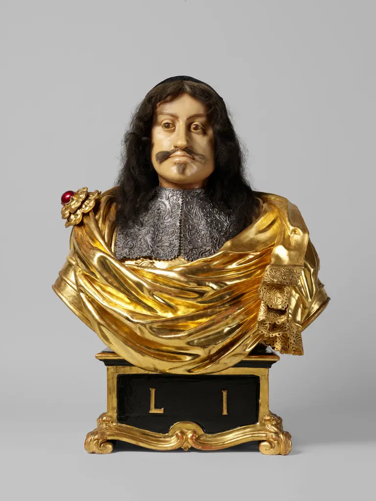 Eine goldene Büste eines Herrschers mit langen Haaren und einem Schnurrbart. Er trägt eine kunstvoll verzierte Brustplatte und eine prächtige goldene Drapierung. Unter der Büste befindet sich die Inschrift 