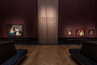 Innenansicht eines Ausstellungsraums mit dunklen Wänden. An den Wänden hängen mehrere Gemälde, während in der Mitte ein Vorhang mit der Aufschrift „Michelina Wautier invenit et fecit“ zu sehen ist. Zwei Bänke sind im Raum platziert.