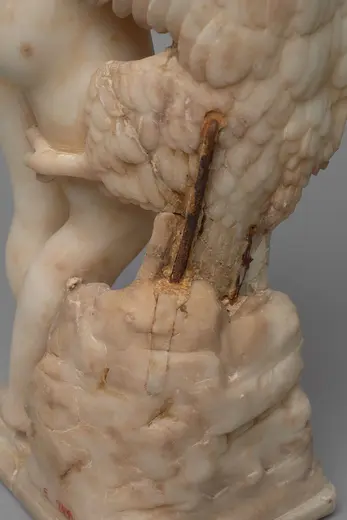 Die Skulptur zeigt einen nackten, jugendlichen menschlichen Körper, der auf einem Block mit einem kunstvoll gestalteten Flügel sitzt. Der Flügel wirkt detailliert und ist aus einem hellen Material gefertigt, während die Basis der Statue rau und bruchstückhaft erscheint.