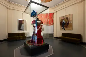 In einem Ausstellungsraum steht unter Glas ein prächtiges rotes Kleid mit blauen Elementen und aufwendigen Verzierungen. Um das Kleid herum sind zwei grüne Bänke und Kunstwerke an den Wänden zu sehen, die die kulturelle Bedeutung der Kleidung betonen.