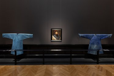 Im Bild sind zwei blaue Kleidungsteile auf einem Tisch zu sehen, die auf Wänden hängen. In der Mitte hängt ein Porträt, das einen Mann mit Bart zeigt. Der Hintergrund ist dunkel, die Umgebung wirkt wie ein Ausstellungsraum.