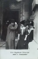 Eine historische Schwarz-Weiß-Fotografie vor dem Burgtheater in Wien. Eine Gruppe von Personen steht vor einem Eingang. Ein Mann mit Zylinder spricht mit einer elegant gekleideten Frau, während andere Damen in historischer Kleidung im Hintergrund stehen.