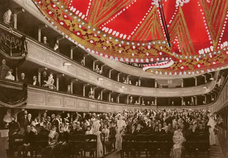 Ein historisches Theaterinnere mit mehreren Ebenen von Zuschauern, die elegant gekleidet sind. Im Vordergrund sitzen Personen auf Stühlen, während im Hintergrund weitere Zuschauer stehen. Eine prunkvolle Decke mit rotem und goldenem Design dominiert die Szene.