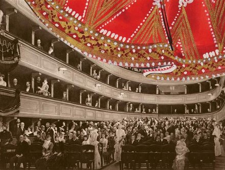 Ein historisches Theaterinnere mit mehreren Ebenen von Zuschauern, die elegant gekleidet sind. Im Vordergrund sitzen Personen auf Stühlen, während im Hintergrund weitere Zuschauer stehen. Eine prunkvolle Decke mit rotem und goldenem Design dominiert die Szene.