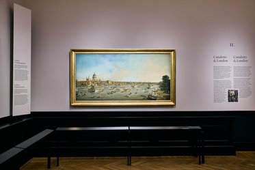 Ein Gemälde zeigt eine Hafenansicht mit Booten und der Stadt im Hintergrund. Das Bild ist in einem goldenen Rahmen gefasst. Neben dem Gemälde hängen Informationen über den Künstler und das Werk an der Wand. Der Raum hat eine moderne Ausstellungsgestaltung.