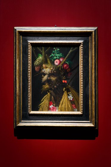 Ein Gemälde in einem goldenen Rahmen zeigt eine maskenartige Figur, die aus verschiedenen Früchten und Pflanzen besteht. Die Figur hat eine natürliche, organische Erscheinung und ist vor einem roten Hintergrund platziert.