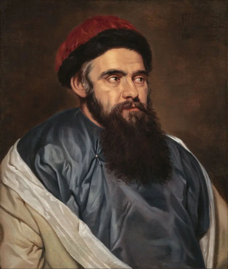 A close-up portrait of a man with a long beard and a thoughtful expression. He is wearing a dark blue garment with a white draped shawl and a red cap. The background is dark, emphasizing the man's face and attire.