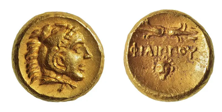Eine goldene Münze, die auf einer Seite das Profil eines Mannes mit lockigem Haar zeigt, möglicherweise eine historische Figur. Die andere Seite trägt den griechischen Namen „ΦΙΛΙΠΠΟΥ“, umgeben von einem dekorativen Muster.