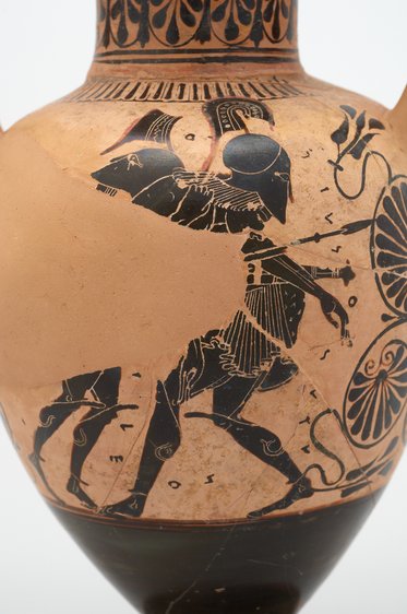 Ein antiker griechischer Krug zeigt einen Krieger in Rüstung mit einem Helm und einem Schild. Er steht in einer kampfbereiten Pose, während er einen Speer hält. Im Hintergrund sind dekorative Muster und Details zu sehen, die die mythologische oder militärische Thematik unterstreichen.