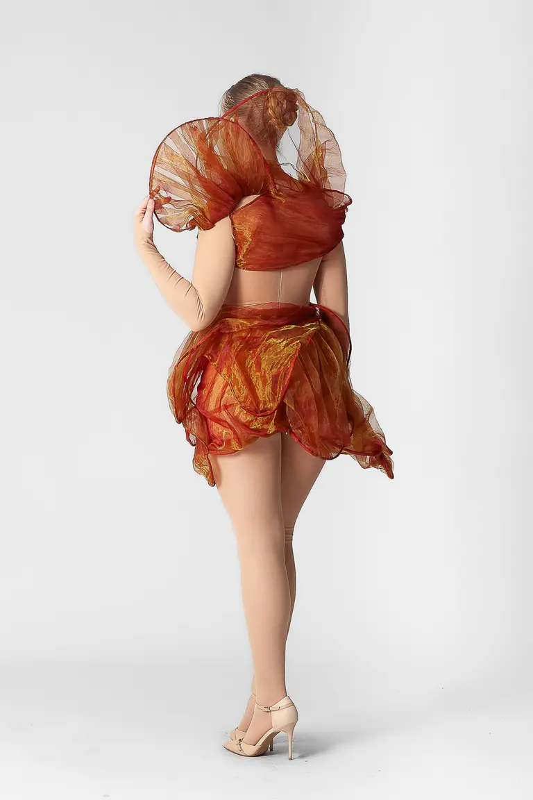 A person is seen from the back wearing a vibrant, ruffled outfit with sheer, flowing fabric in shades of orange and red. The design features large, circular elements around the shoulders and a playful skirt. The model stands on a plain white background, showcasing the outfit's movement.