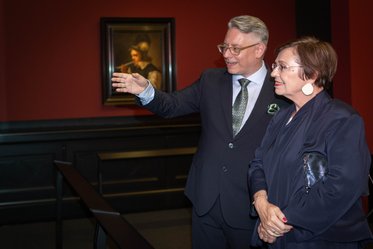 Ein Mann im Anzug zeigt auf ein Kunstwerk an der Wand, während eine Frau in eleganter Kleidung ihn aufmerksam anhört. Der Hintergrund ist rot gestrichen und das Kunstwerk ist ein Porträt.