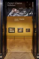 Eingang zu einer Ausstellung über den Maler Pieter Claesz, die seine Stillleben präsentiert. An der Wand sind mehrere Gemälde zu sehen, während der Raum in warmen Gelbtönen gestaltet ist. Ein großes Banner mit dem Namen des Künstlers und dem Thema der Ausstellung hängt über dem Eingang.