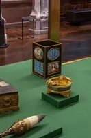 Auf einem grünen Tisch sind drei Objekte zu sehen: Ein quadratischer, verziierter Behälter mit Bildern, eine runde, goldene Dose und ein Pinsel mit einem breiten Borstenkopf. Die Objekte stammen aus einer historischen Sammlung und sind vor einer Glasvitrine ausgestellt.