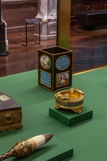 Auf einem grünen Tisch sind drei Objekte zu sehen: Ein quadratischer, verziierter Behälter mit Bildern, eine runde, goldene Dose und ein Pinsel mit einem breiten Borstenkopf. Die Objekte stammen aus einer historischen Sammlung und sind vor einer Glasvitrine ausgestellt.