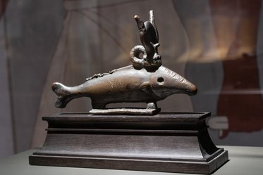 Bronzeskulptur eines Fisches mit einem Horn und einer Spirale als Kopfteil, auf einem rechteckigen Sockel aus Holz platziert. Der Fisch hat eine glatte Oberfläche und ist detailliert gearbeitet. Hintergrund unscharf, mit angedeuteten menschlichen Figuren.