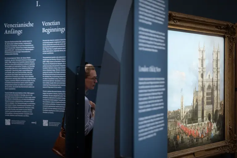 Eine Person steht in einer Ausstellung vor einem Gemälde, das eine historische Szene mit einer Kathedrale und einer Menschenmenge zeigt. Im Hintergrund sind Schautafeln mit Texten zu sehen, die Informationen über die venezianische Kunst vermitteln.
