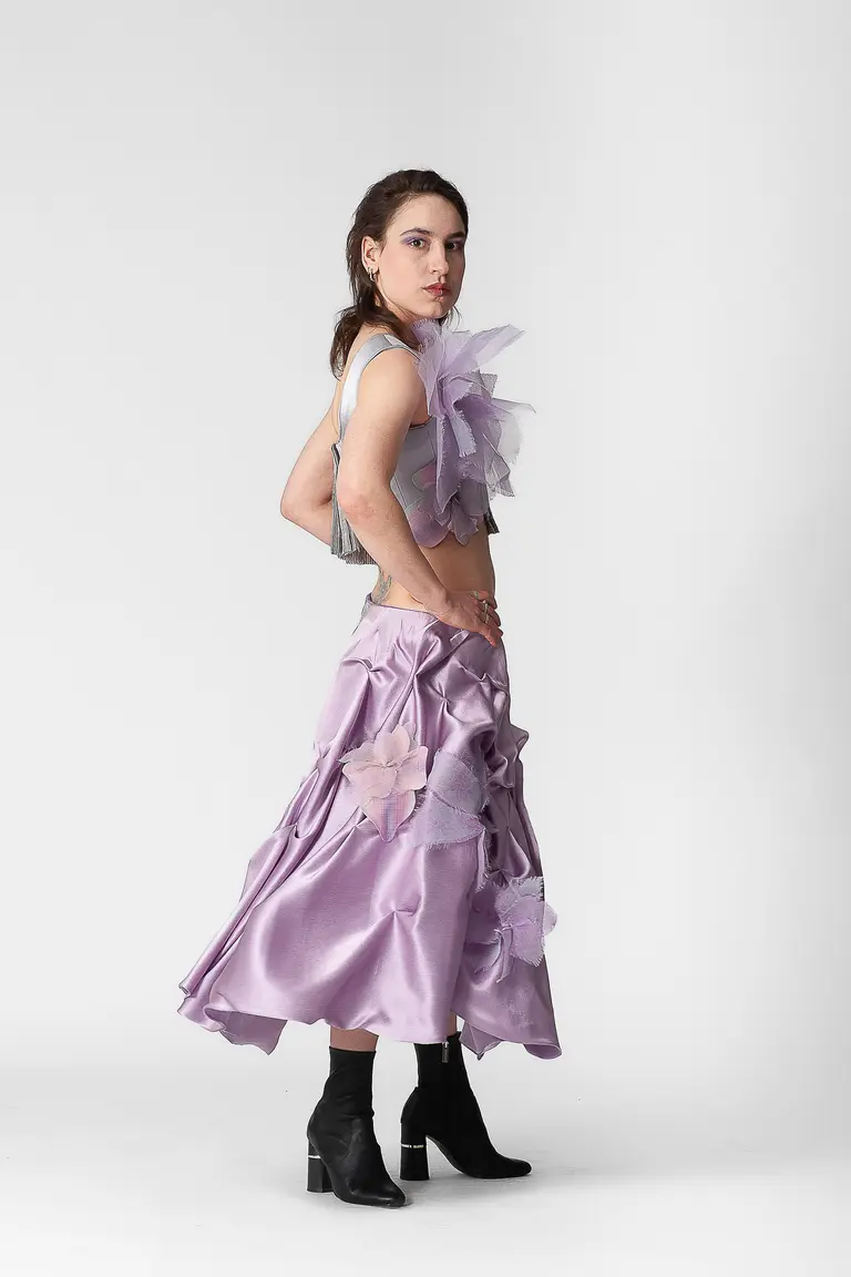 A model poses sideways, wearing a unique outfit featuring a cropped top with layered, translucent elements and a long, flowing lavender skirt adorned with floral appliqués. She wears black ankle boots and has her hair styled down, creating a striking and artistic look against a plain background.